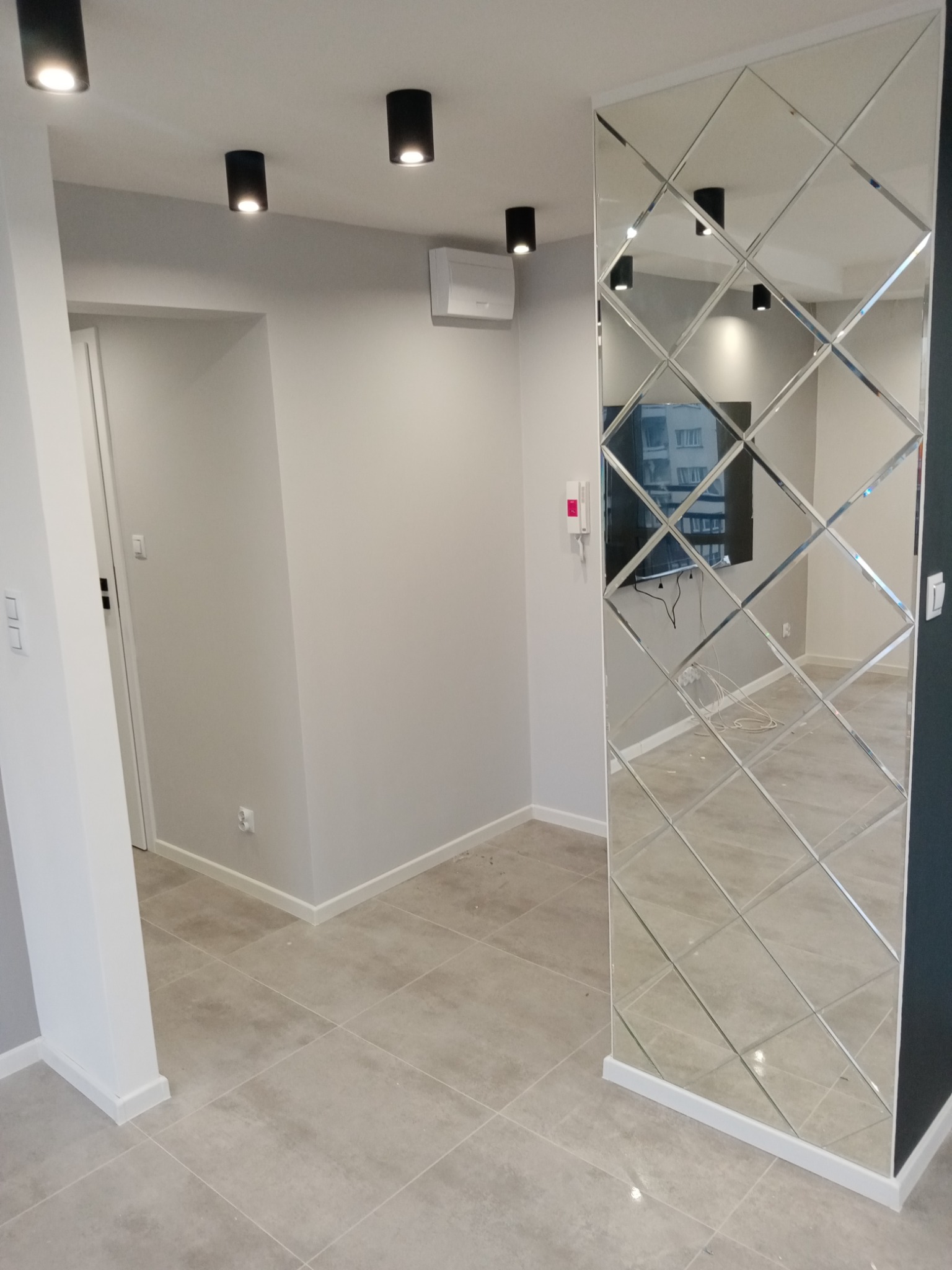 Nowoczesny salon z szarą podłogą z płytek, lustrzaną ścianą z romboidalnych paneli, białymi ścianami i czarnymi lampami sufitowymi