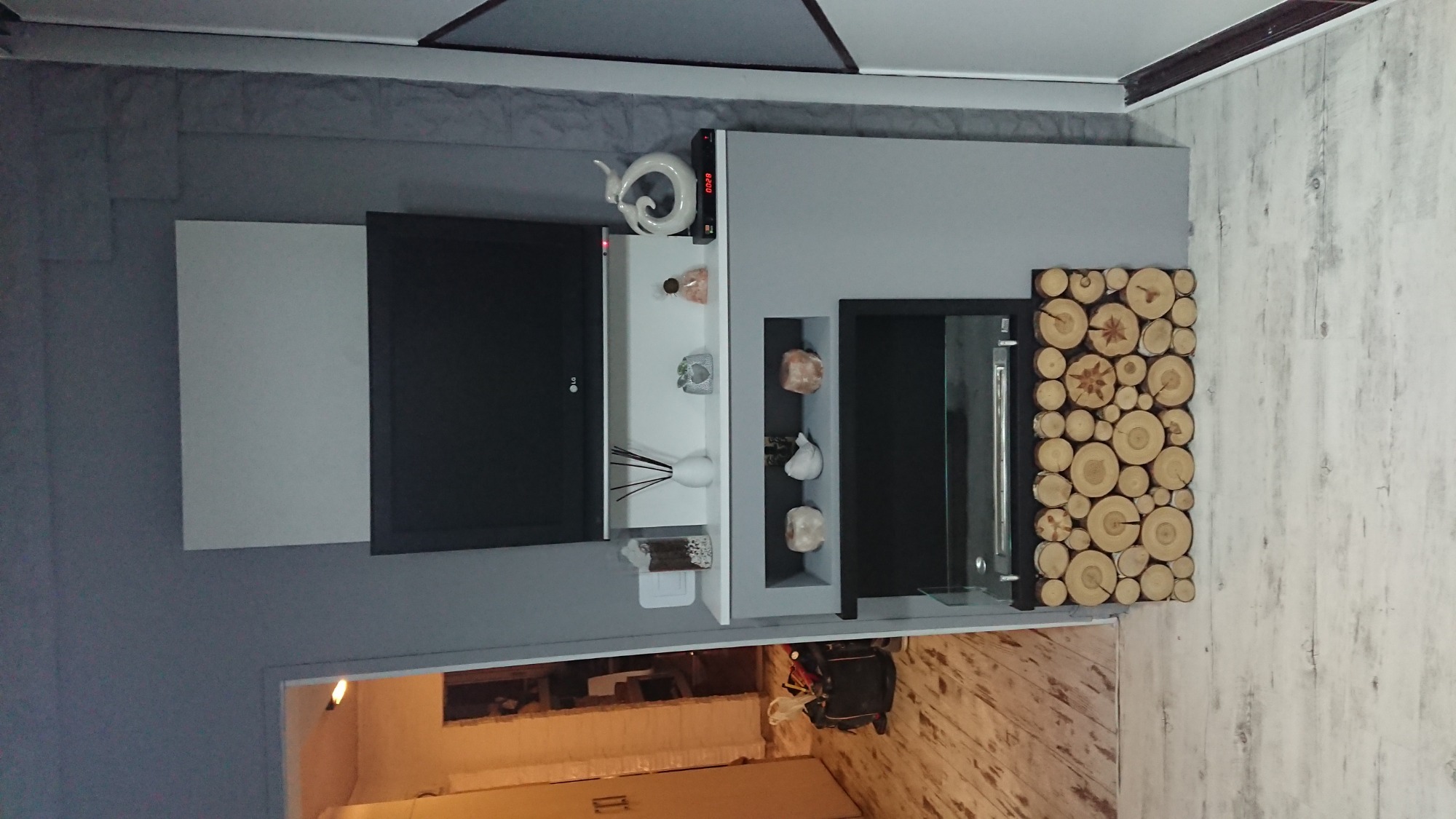 Szara ściana z telewizorem, półkami dekoracyjnymi i panelem z plastrów drewna, widok na remontowany pokój.