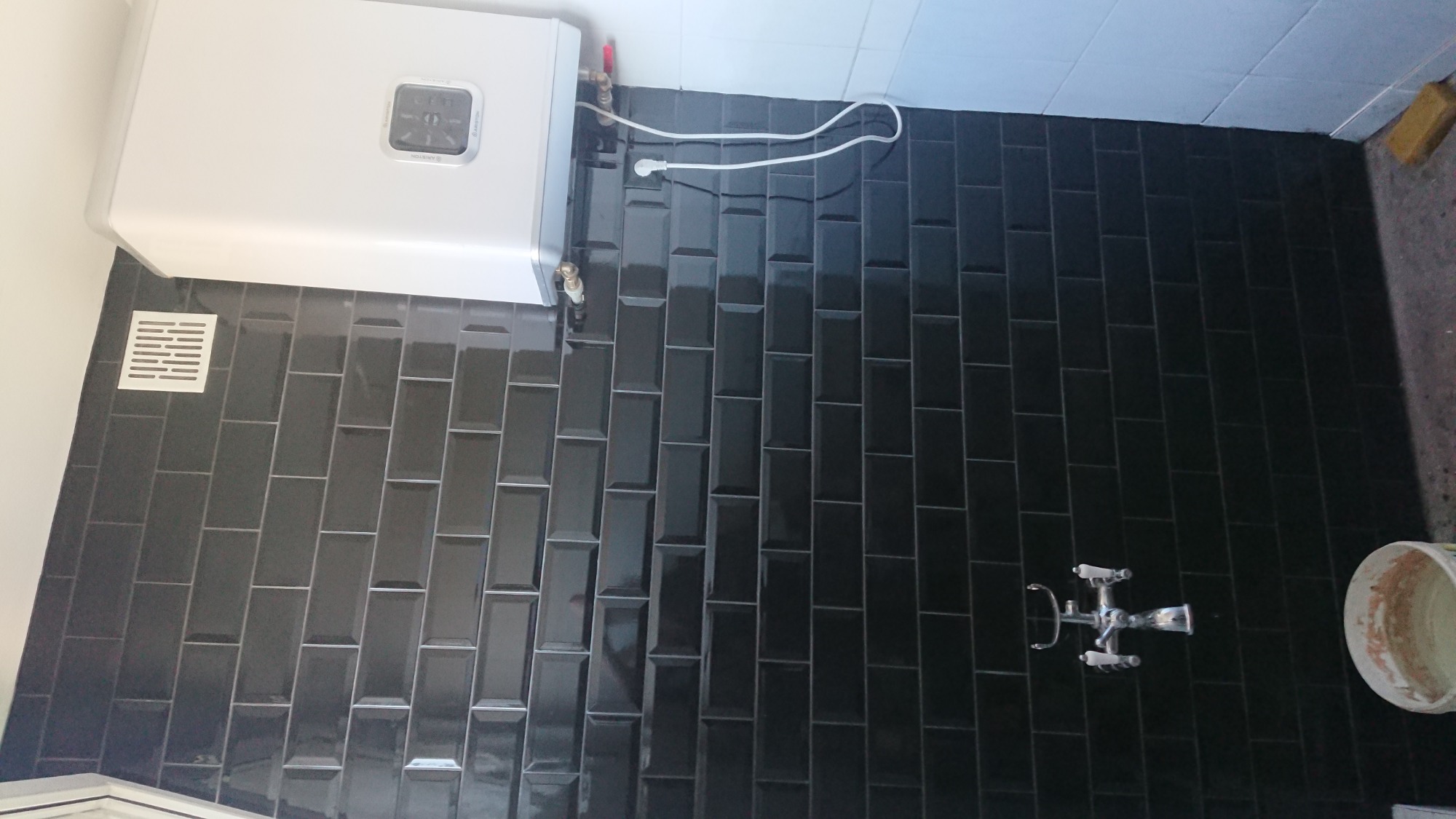 Czarna, błyszcząca glazura w łazience, z widocznym bojlerem elektrycznym, kratką wentylacyjną i baterią umywalkową na ścianie, pod lekkim kątem.