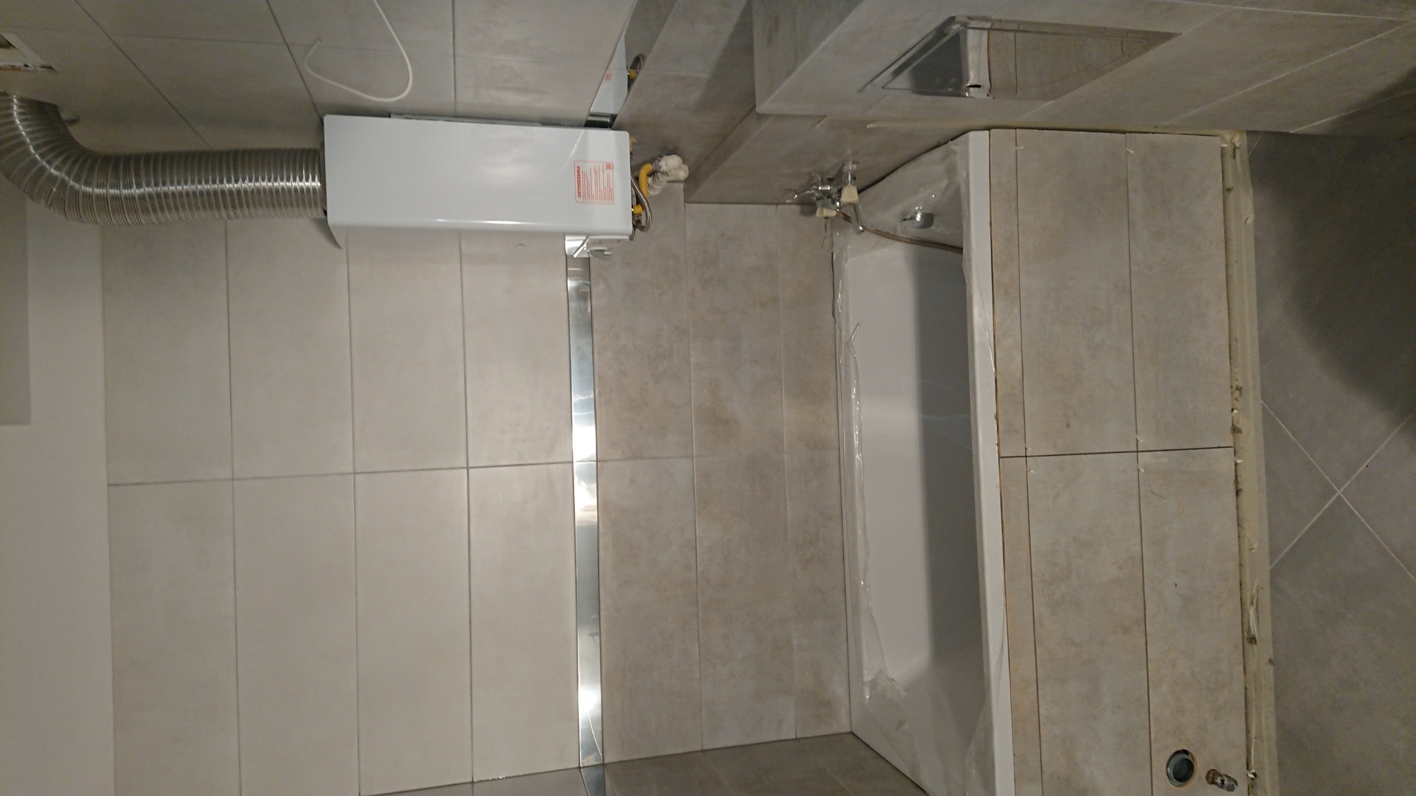 Montaż bojlera z podłączeniem wentylacyjnym w łazience z jasnymi płytkami, widoczna wanna zabezpieczona folią.