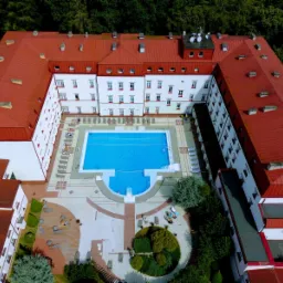 Widok z lotu ptaka na hotel z czerwonym dachem i basenem na dziedzińcu, otoczonym leżakami i zielenią.