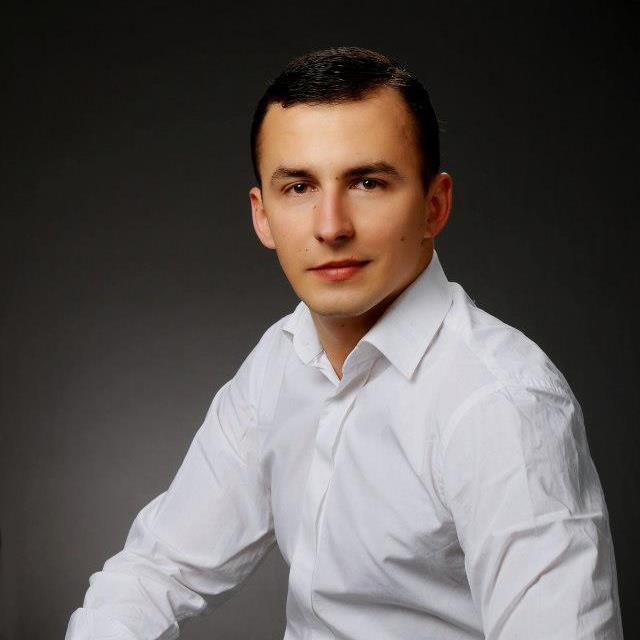 Portret młodego mężczyzny w białej koszuli na szarym tle, uśmiechający się lekko do kamery.