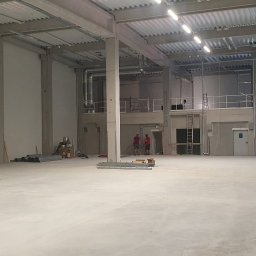 MSARCH Krzysztof Malczewski - Wnętrze nowej hali produkcyjnej w trakcie wykańczania, widoczne betonowe słupy, metalowa konstrukcja dachu, instalacje wentylacyjne, oraz pracownicy przy pracy.