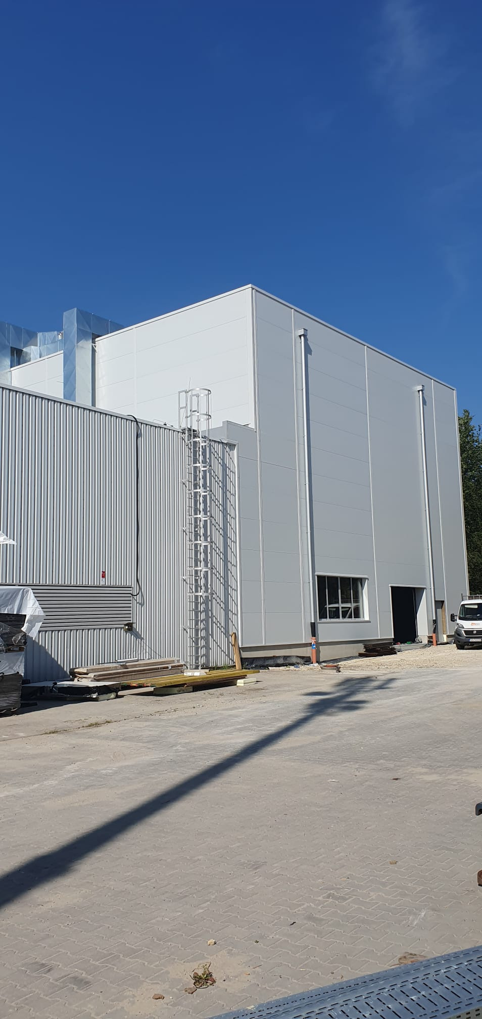 Nowoczesny budynek usługowy o szarej elewacji z blachy trapezowej, z metalową drabinką na ścianie, widoczny z zewnątrz w słoneczny dzień, z zaparkowanym białym samochodem dostawczym w tle.