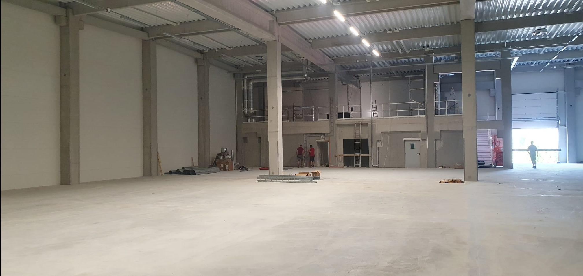 Wnętrze nowej hali produkcyjnej w trakcie wykańczania, widoczne betonowe słupy, metalowa konstrukcja dachu, instalacje wentylacyjne, oraz pracownicy przy pracy.