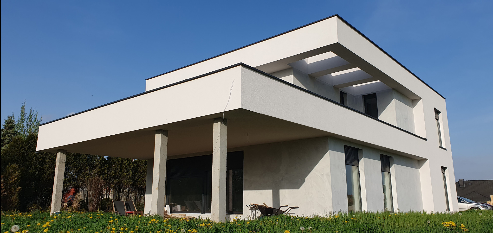 Nowoczesny, dwukondygnacyjny dom z płaskim dachem, białymi ścianami i betonowymi kolumnami podtrzymującymi balkon, otoczony zielonym trawnikiem z mleczami, w tle drzewa i błękitne niebo.