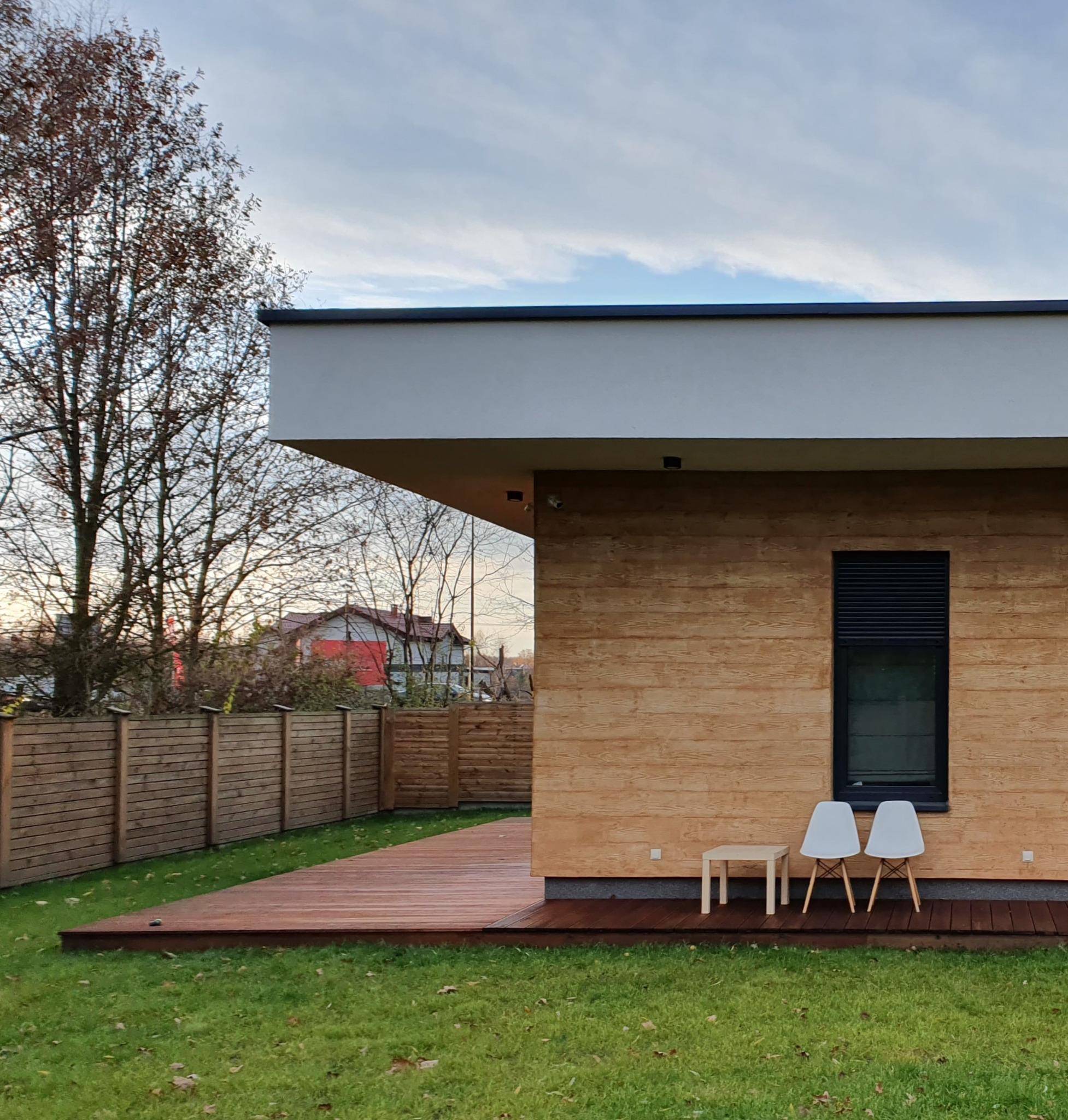 Nowoczesny dom jednorodzinny z drewnianą elewacją, tarasem z desek i dwoma białymi krzesłami przed oknem z żaluzjami, otoczony trawnikiem i drewnianym ogrodzeniem w jesiennej scenerii.