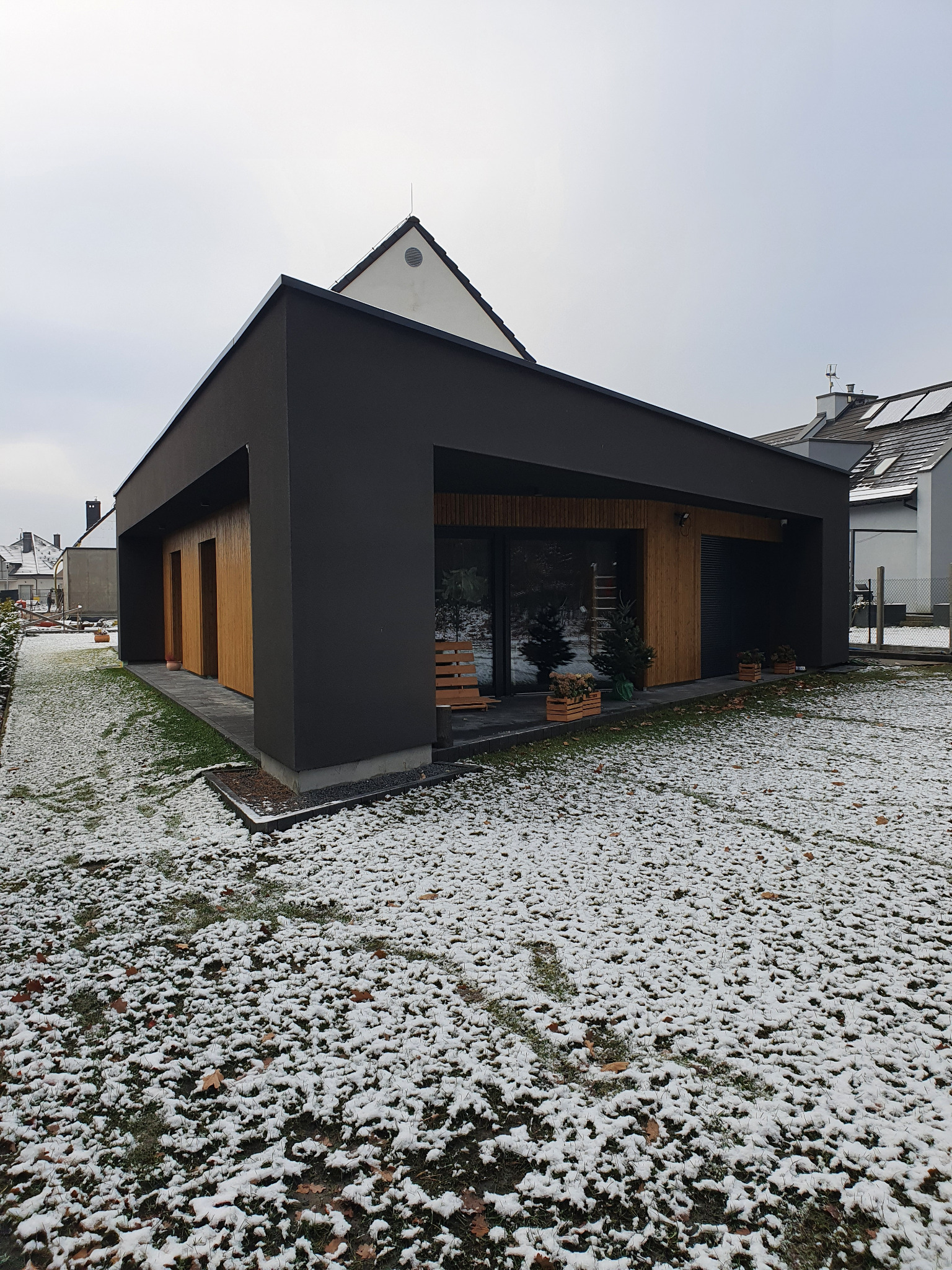 Nowoczesny, parterowy dom w ciemnej kolorystyce z elementami drewna, otoczony trawnikiem pokrytym częściowo śniegiem. W tle widoczne inne domy.