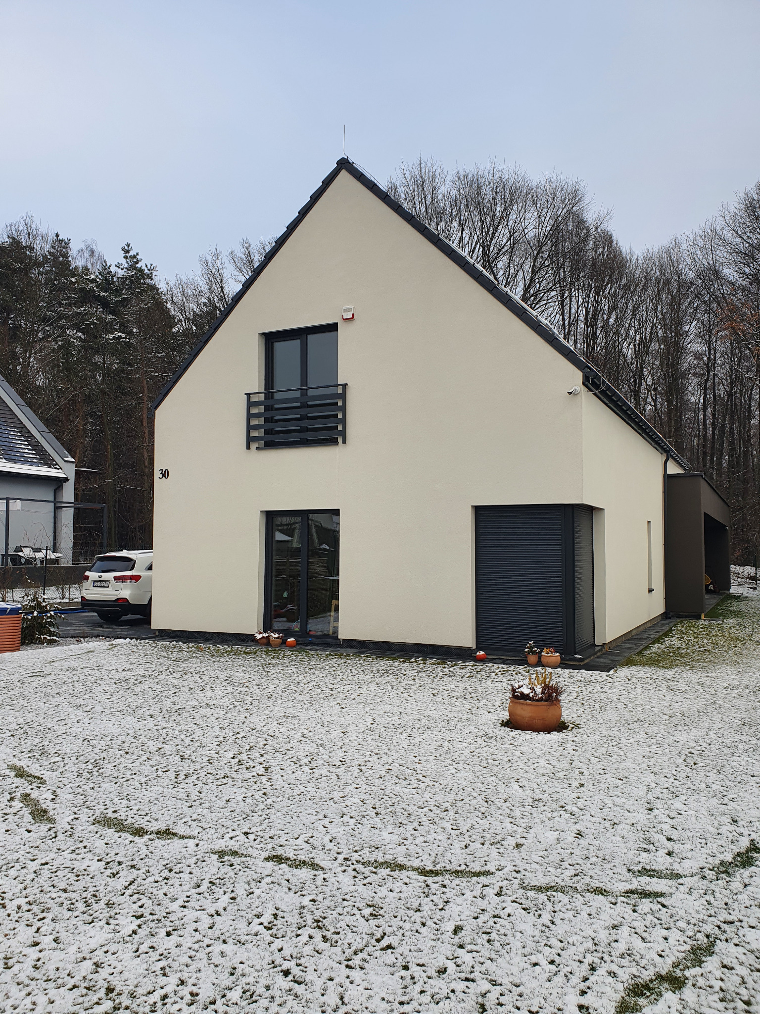 Nowoczesny, dwukondygnacyjny dom z płaskim dachem i numerem 30, kremową elewacją, czarnymi ramami okien i drzwi oraz garażem z roletą, otoczony trawnikiem pokrytym śniegiem i drzewami bez liści.