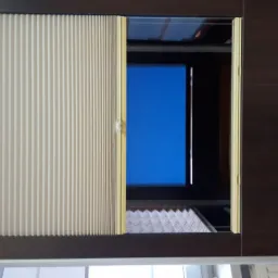 Fragment okna z beżową plisowaną osłoną częściowo zasłoniętą i ciemnoszarą pionową osłoną po prawej stronie, widok na błękitne niebo przez otwartą przestrzeń.