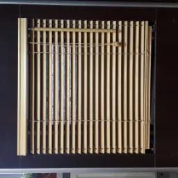 Beżowe pionowe listwy w oknie, obok biała tkanina i brązowe wąskie panele, widoczne mocowania i sznurki.