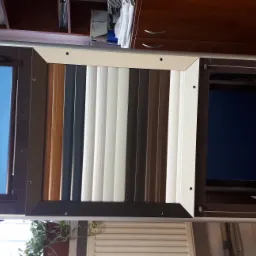 Ekspozycja próbek różnych kolorów i materiałów systemów osłonowych okien, w tym pionowe lamele i zwinięte ekrany, na tle mebli biurowych.