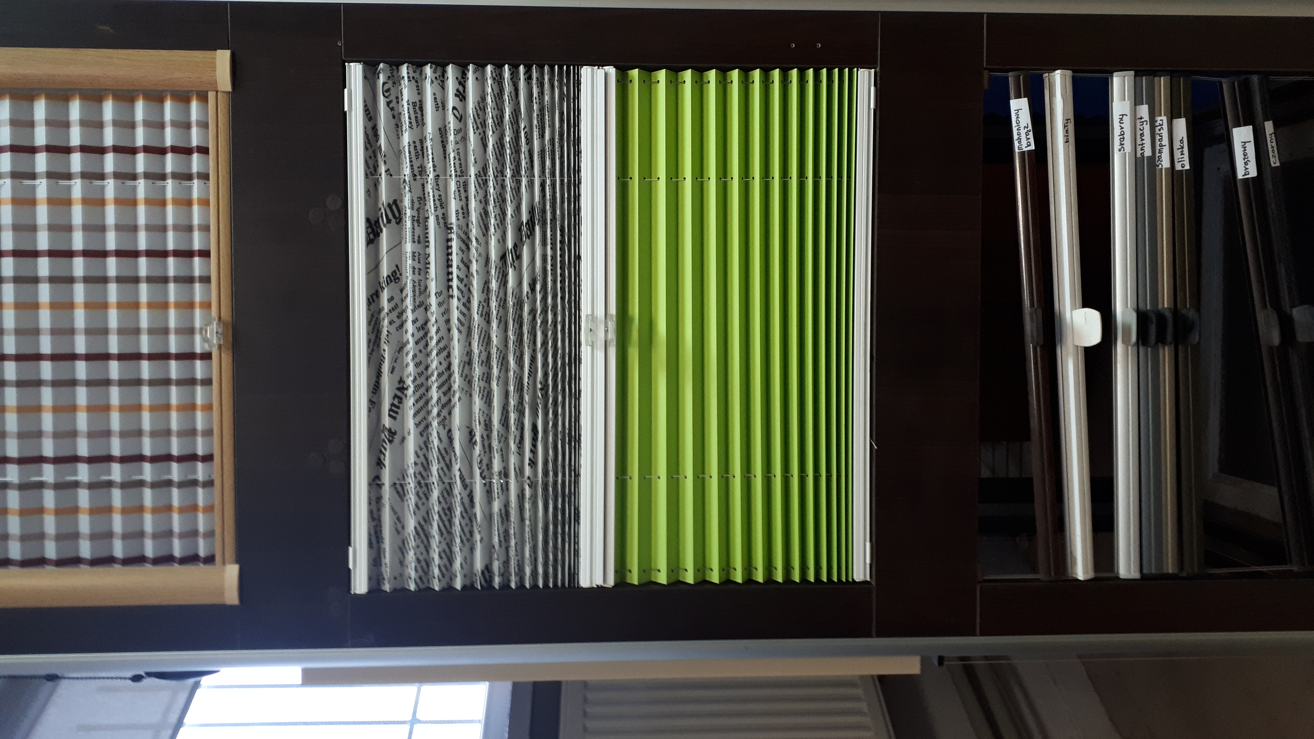 Ekspozycja próbek plisowanych przesłon okiennych: krata, wzór gazetowy i jednolity zielony, obok profile w różnych kolorach z etykietami.