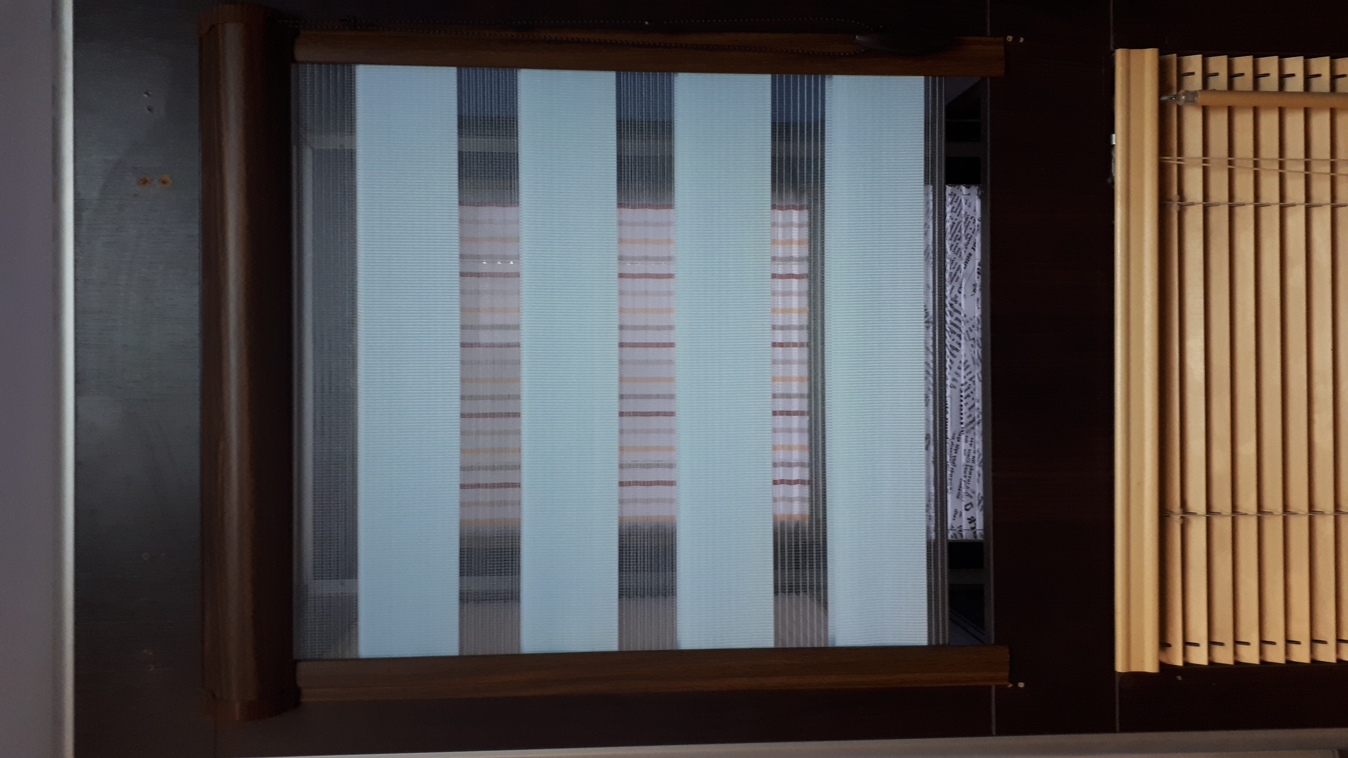 Okno z zamontowanymi pionowymi panelami tkaninowymi w odcieniach bieli i szarości, z widocznym wzorem w paski i drewnianą ramą, obok bambusowej osłony okiennej.