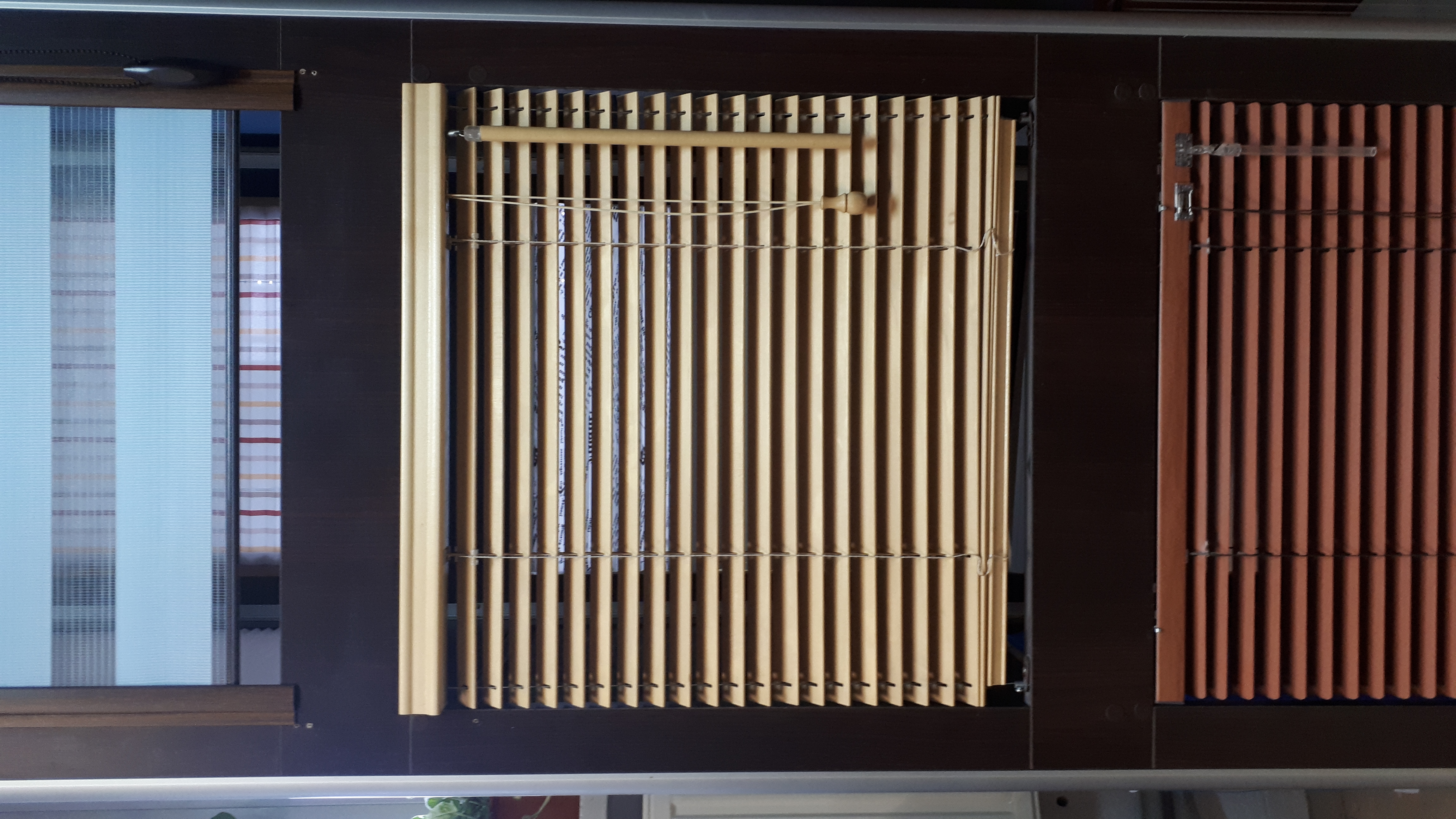 Beżowe pionowe listwy w oknie, obok biała tkanina i brązowe wąskie panele, widoczne mocowania i sznurki.