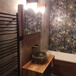 Nowoczesna łazienka z umywalką z kamienia, drewnianą szafką, grzejnikiem drabinkowym i ścianą w tropikalne wzory.