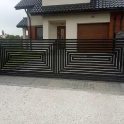 Nowoczesna, czarna brama wjazdowa z geometrycznym wzorem prostokątów, osadzona na kamiennych słupkach, na tle domu z brązową bramą garażową i szarą dachówką.