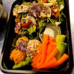 Kompozycja sałatkowa w czarnym pojemniku: sałata, pomidory, ogórki, ser pleśniowy, pestki, kiełki, pasta warzywna, pasta z tuńczyka, marchewka, sos w plastikowym pojemniczku.