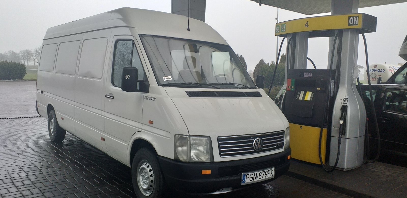 Biały bus Volkswagen LT35 na stacji benzynowej, widoczny dystrybutor paliwa i mokra kostka brukowa.