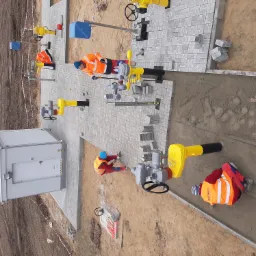 Montaż instalacji gazowej i układanie szarej kostki brukowej wokół infrastruktury, widok z góry na plac budowy z pracownikami w odblaskowych kamizelkach.