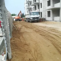 Plac budowy z ciężarówką, koparką Atlas i robotnikami w odblaskowych ubraniach, widoczne elementy konstrukcji budynku i palety z materiałami budowlanymi.