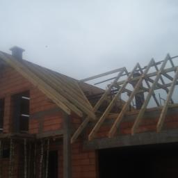 Piotr Jagiełło - Wznoszenie drewnianej konstrukcji dachu na murowanym budynku w trakcie budowy, widoczne elementy więźby dachowej i komin w surowym stanie.