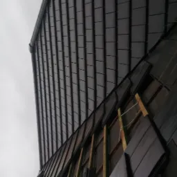 Fragment dachu z ciemnej dachówki podczas montażu, widoczne elementy konstrukcji drewnianej i podszycie, pochmurne niebo w tle.