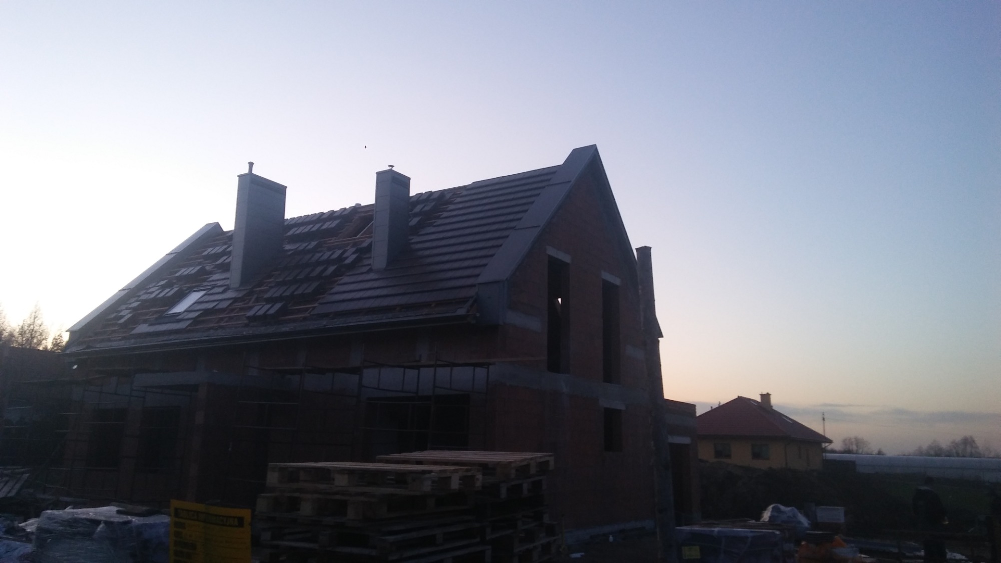 Dom w trakcie krycia dachu czerwoną dachówką ceramiczną z widocznymi kominami, w tle błękitne niebo o zmierzchu.