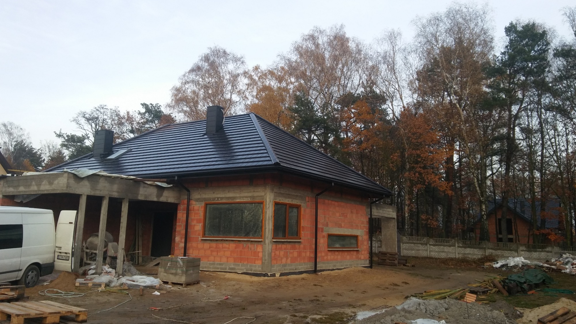 Dom w stanie surowym z nowym, czarnym dachem z dachówki ceramicznej, widoczne kominy i rynny, otoczenie w trakcie budowy, jesienne drzewa w tle.