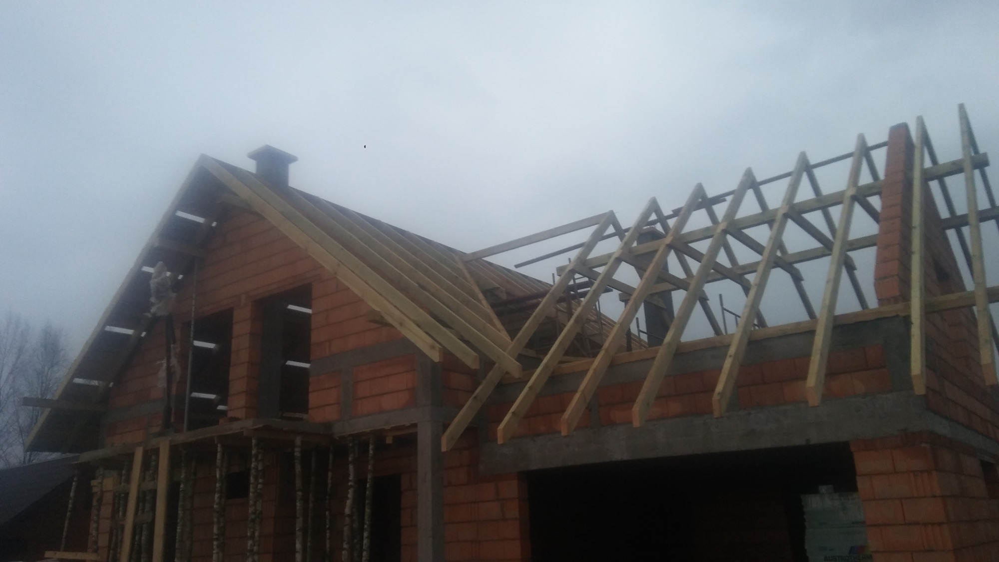 Wznoszenie drewnianej konstrukcji dachu na murowanym budynku w trakcie budowy, widoczne elementy więźby dachowej i komin w surowym stanie.