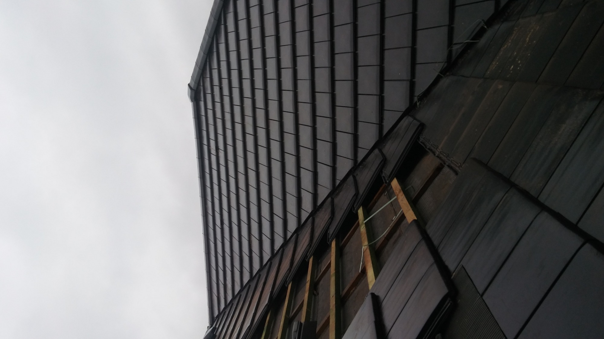 Fragment dachu z ciemnej dachówki podczas montażu, widoczne elementy konstrukcji drewnianej i podszycie, pochmurne niebo w tle.