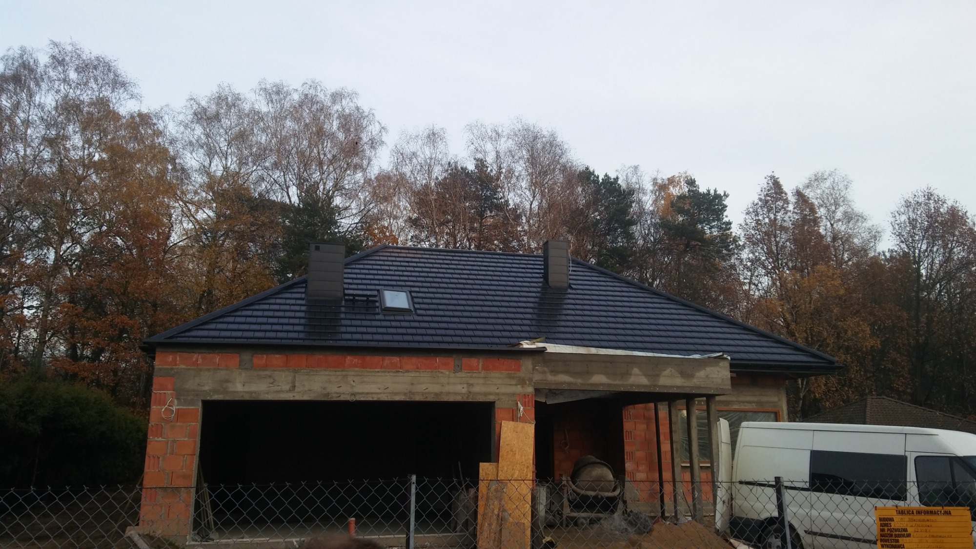 Nowo położony, ciemny dach z dwoma kominami i oknem dachowym na surowym budynku z czerwonej cegły, na tle jesiennych drzew.