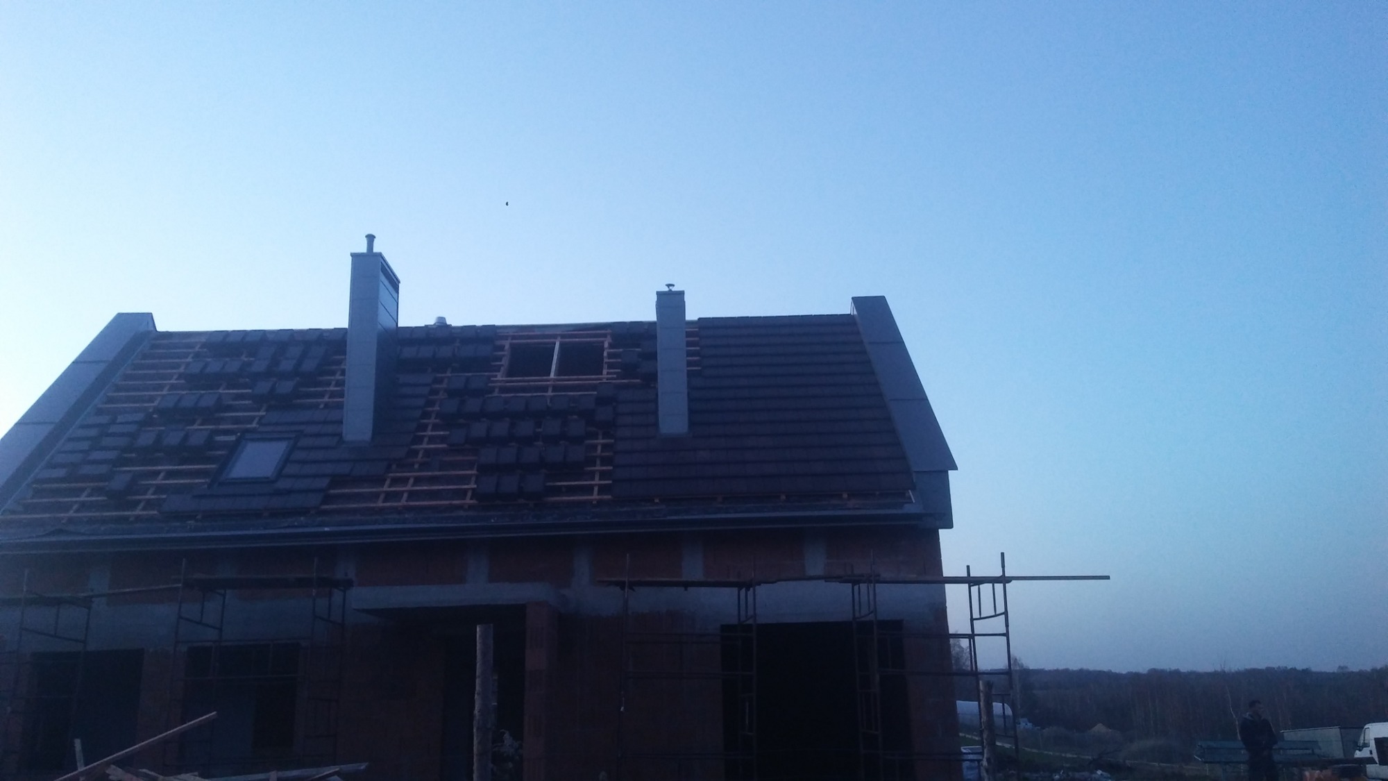 Wznoszony dom jednorodzinny z częściowo pokrytym dachem grafitową dachówką, widoczne kominy i okno dachowe, na pierwszym planie rusztowania.