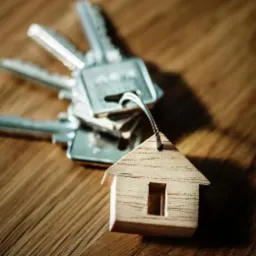 Pęk kluczy z drewnianym breloczkiem w kształcie domu na drewnianym blacie, symbolizujący zarządzanie nieruchomościami.
