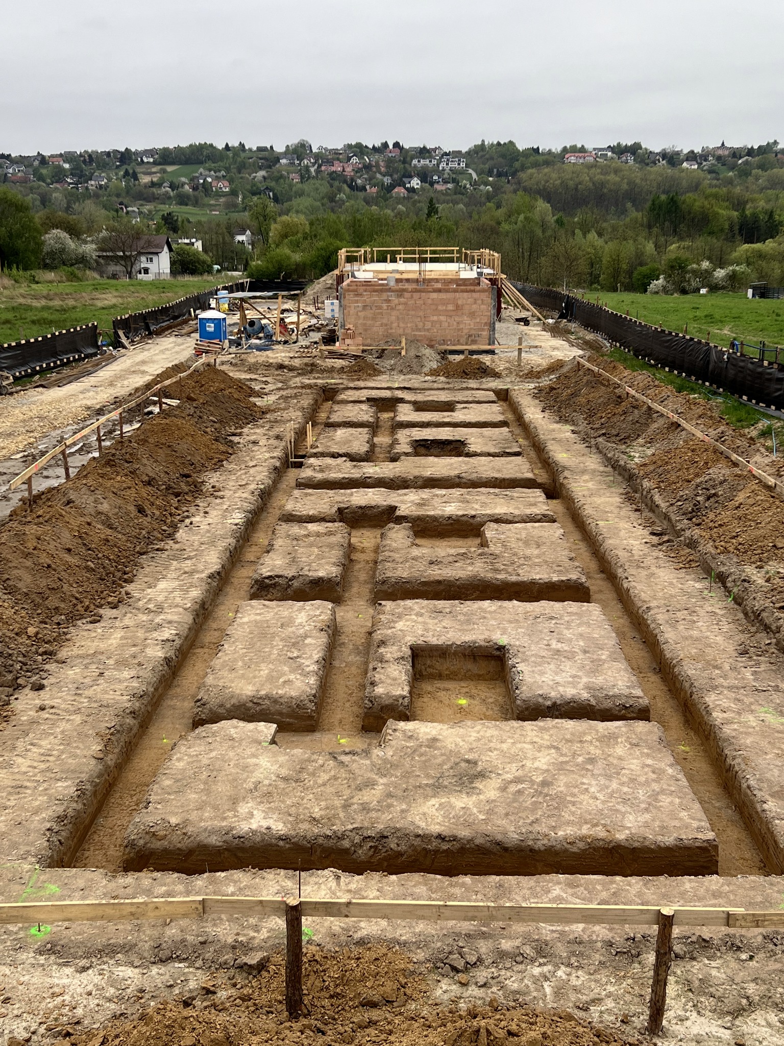 Wykonane wykopy pod fundamenty domu jednorodzinnego, widoczne z góry. W tle zabudowa mieszkalna na wzgórzu i częściowo wymurowane ściany fundamentowe.
