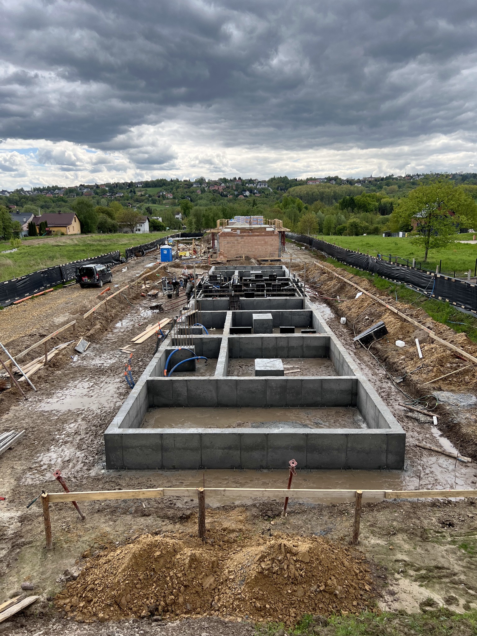 Fundamenty domu w trakcie budowy, widok z góry na zalane betonem ściany fundamentowe, zbrojenia i instalacje. W tle krajobraz z zabudowaniami i pochmurne niebo.