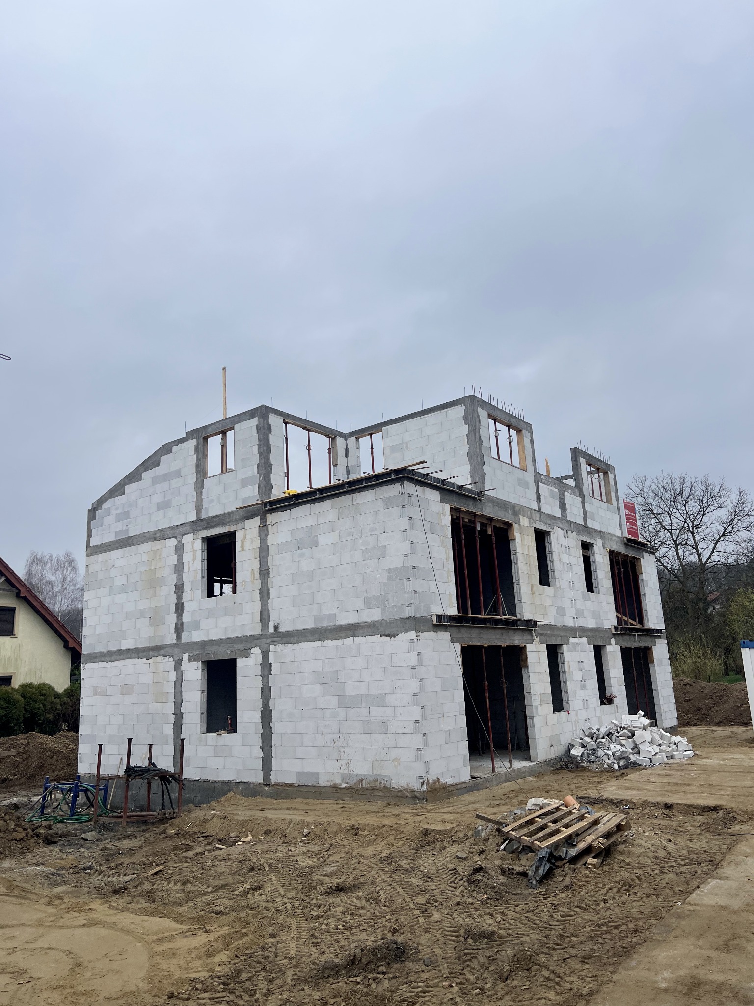Dwukondygnacyjny dom w trakcie budowy z białych bloczków, widoczne otwory okienne i drzwiowe, szare niebo w tle. Na ziemi gruz i paleta.