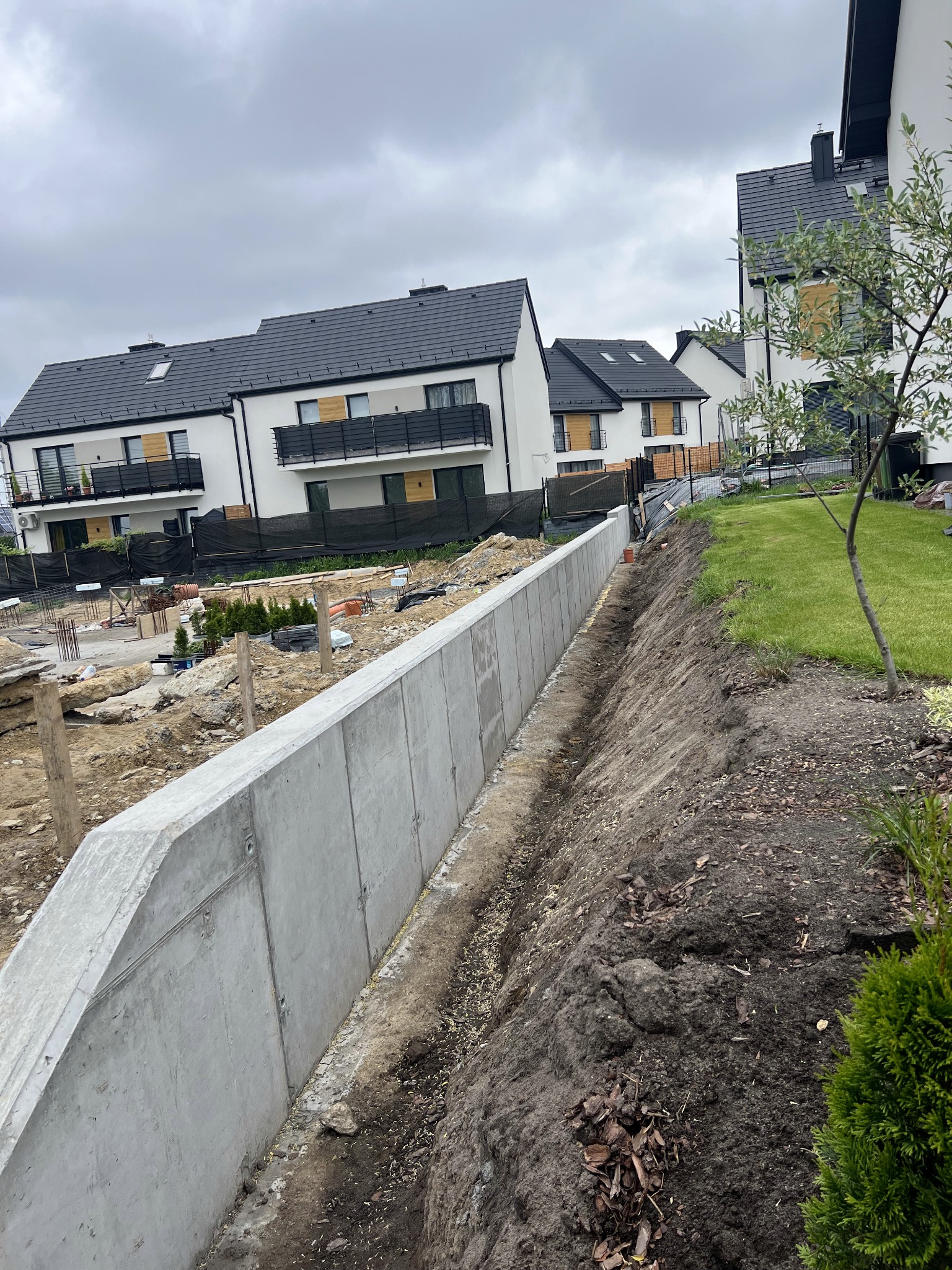 Betonowy mur oporowy w Radziszowie, zabezpieczający skarpę na tle budowanych domów. Widoczne zbrojenia i przygotowany teren pod dalsze prace.