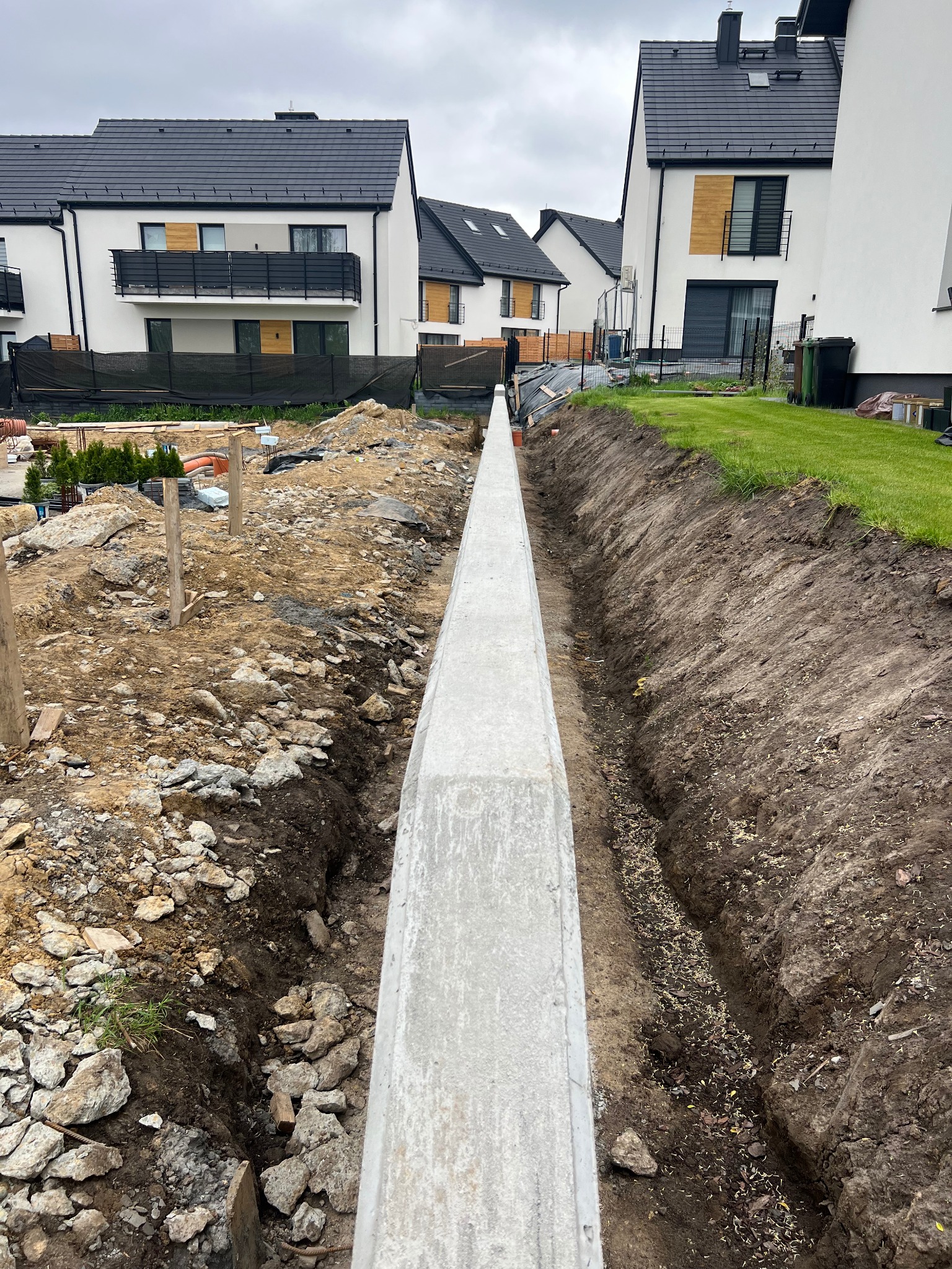 Wykop pod fundament z betonową wylewką na tle budynków w nowoczesnym stylu. Widok z góry na wąski wykop z elementami zbrojenia i gruzu.