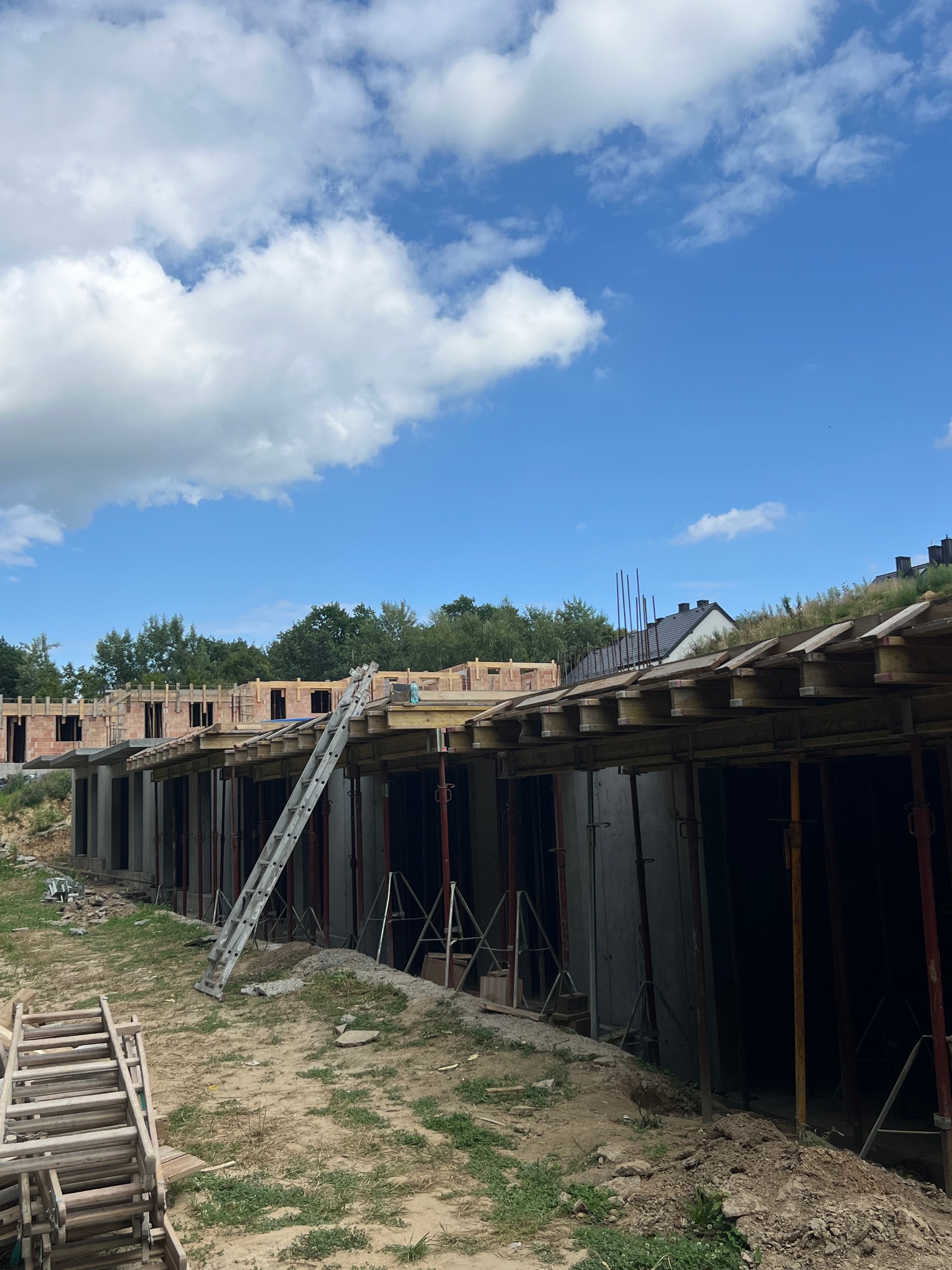 Budowa domu w trakcie: widoczne fundamenty, ściany z czerwonej cegły, drewniane szalunki i drabina oparta o konstrukcję. Błękitne niebo z chmurami w tle.