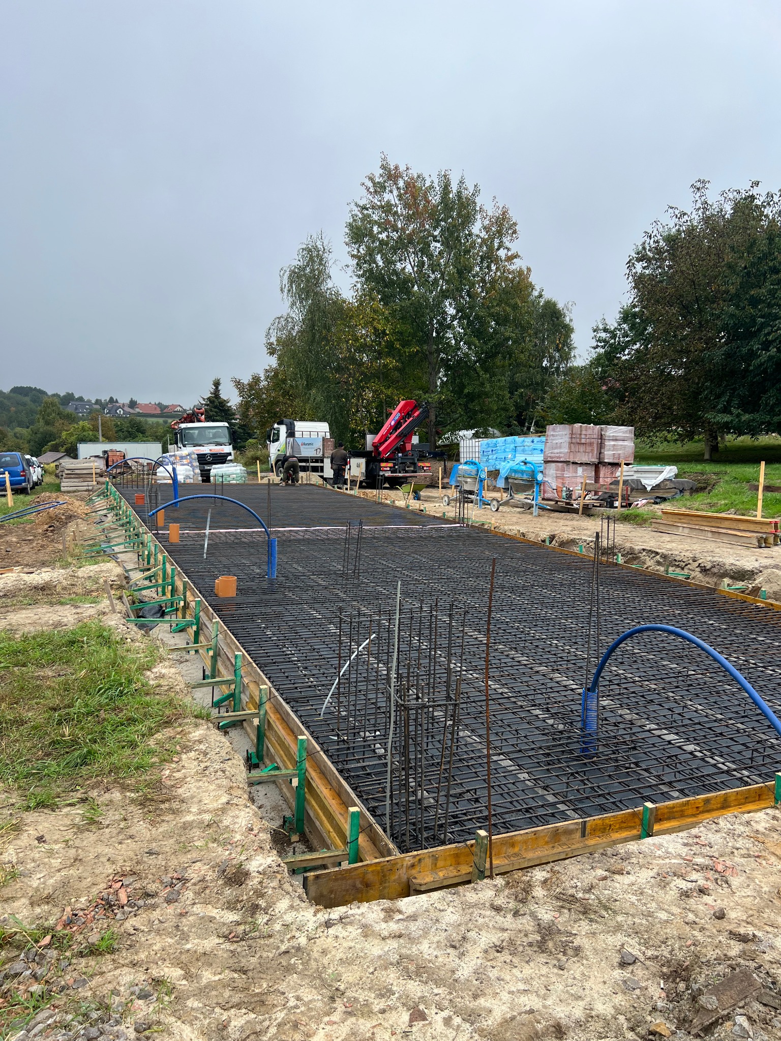 Zbrojenie fundamentów pod budowę domu jednorodzinnego, widoczne szalunki, stalowe pręty, niebieskie rury instalacyjne i sprzęt budowlany w tle. Ujęcie z góry.