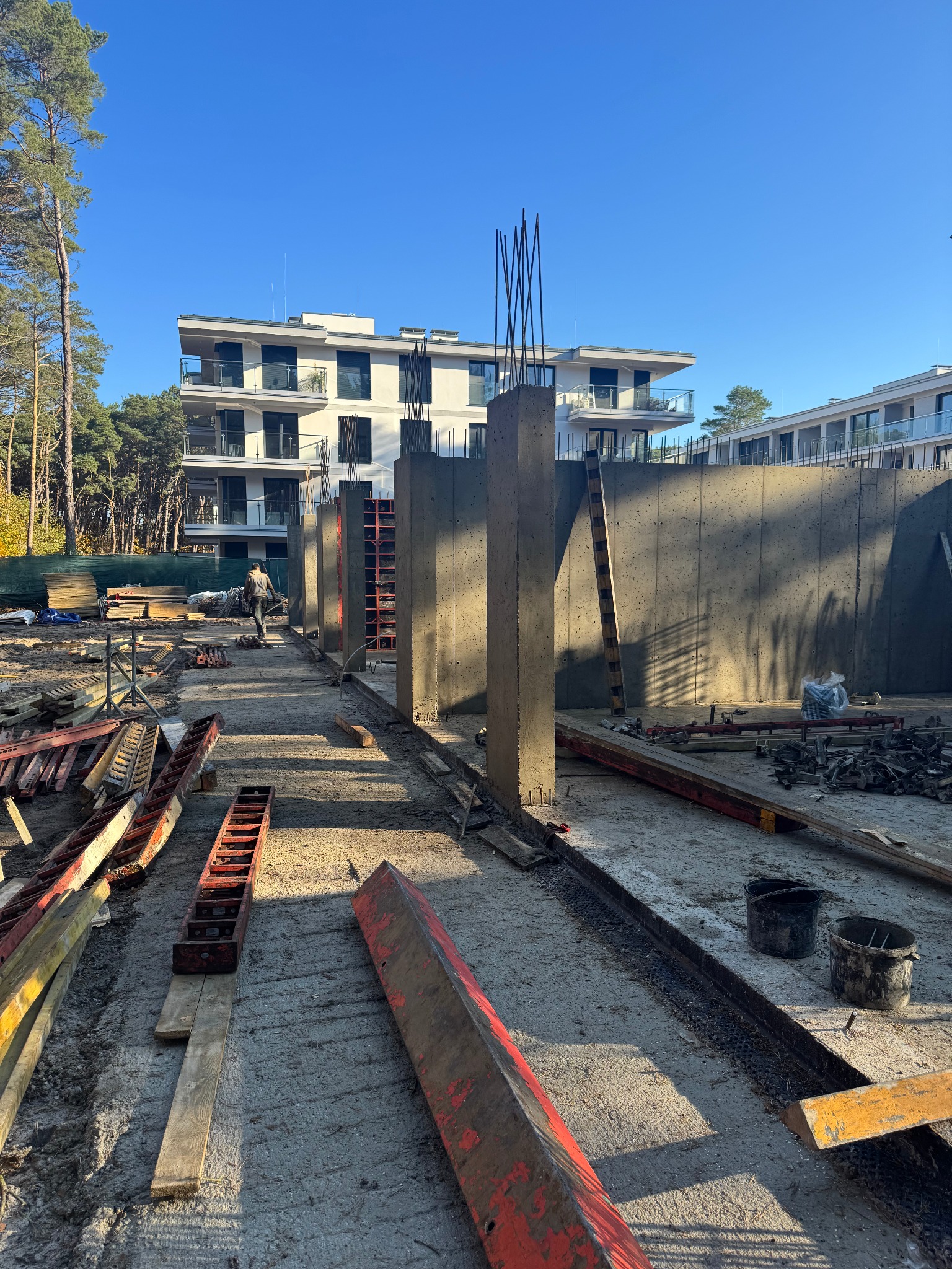 Budowa apartamentowca w trakcie wznoszenia ścian i kolumn z betonu, widoczne szalunki i zbrojenia, w tle drzewa i błękitne niebo.