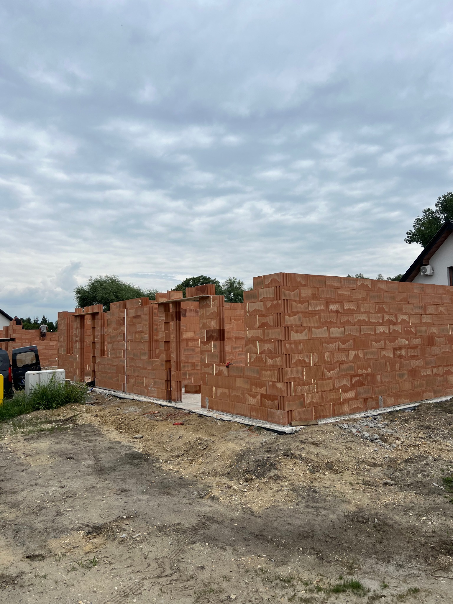 Wznoszenie ścian domu z czerwonej cegły ceramicznej. Widoczne fragmenty ścian z otworami okiennymi i drzwiowymi. Na drugim planie częściowo widoczny samochód i pracownicy.