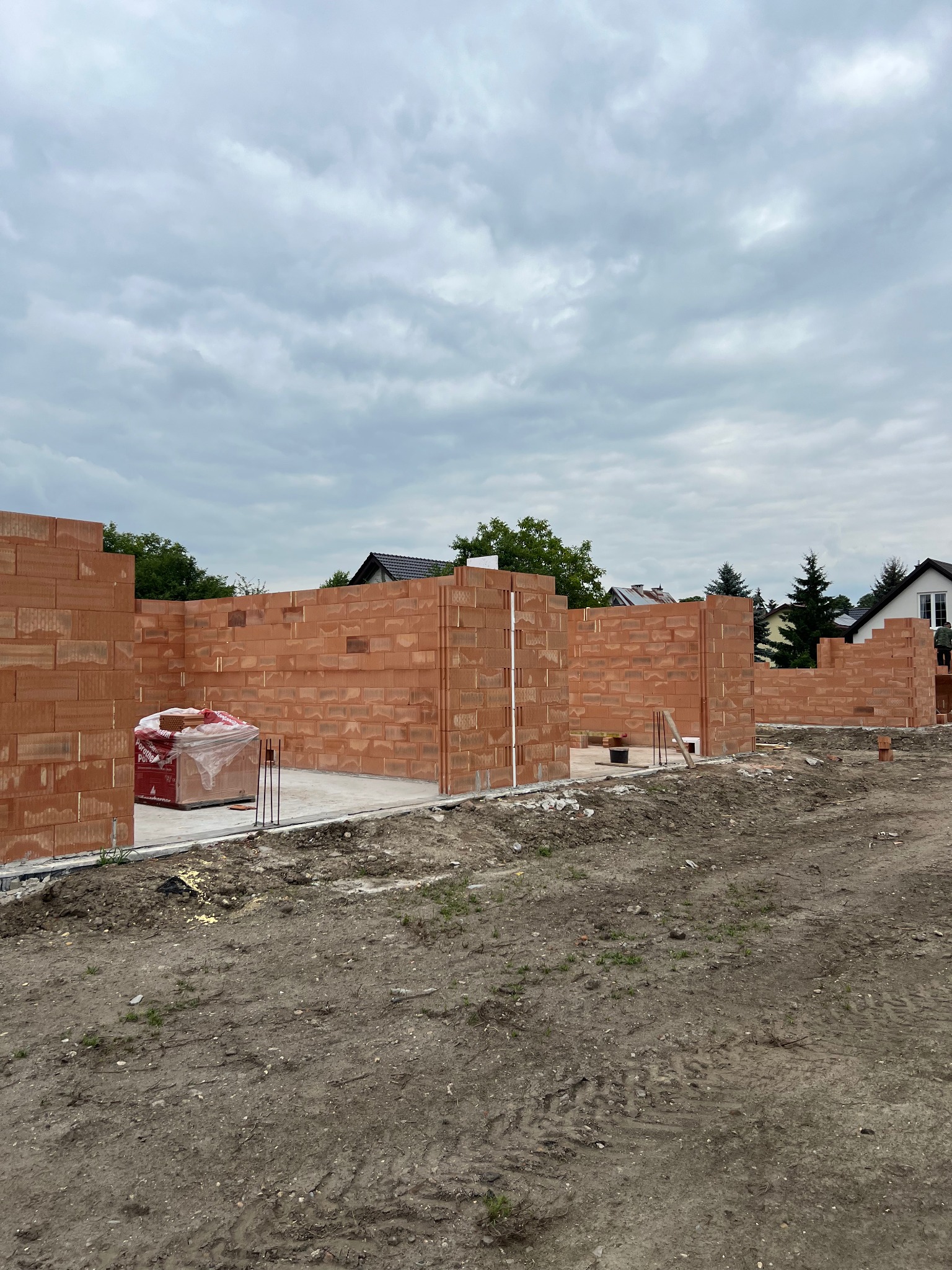 Wznoszone ściany z czerwonej cegły ceramicznej Porotherm na fundamencie. Częściowo zapakowane palety z cegłami. Widok na budowę domu jednorodzinnego w pochmurny dzień.