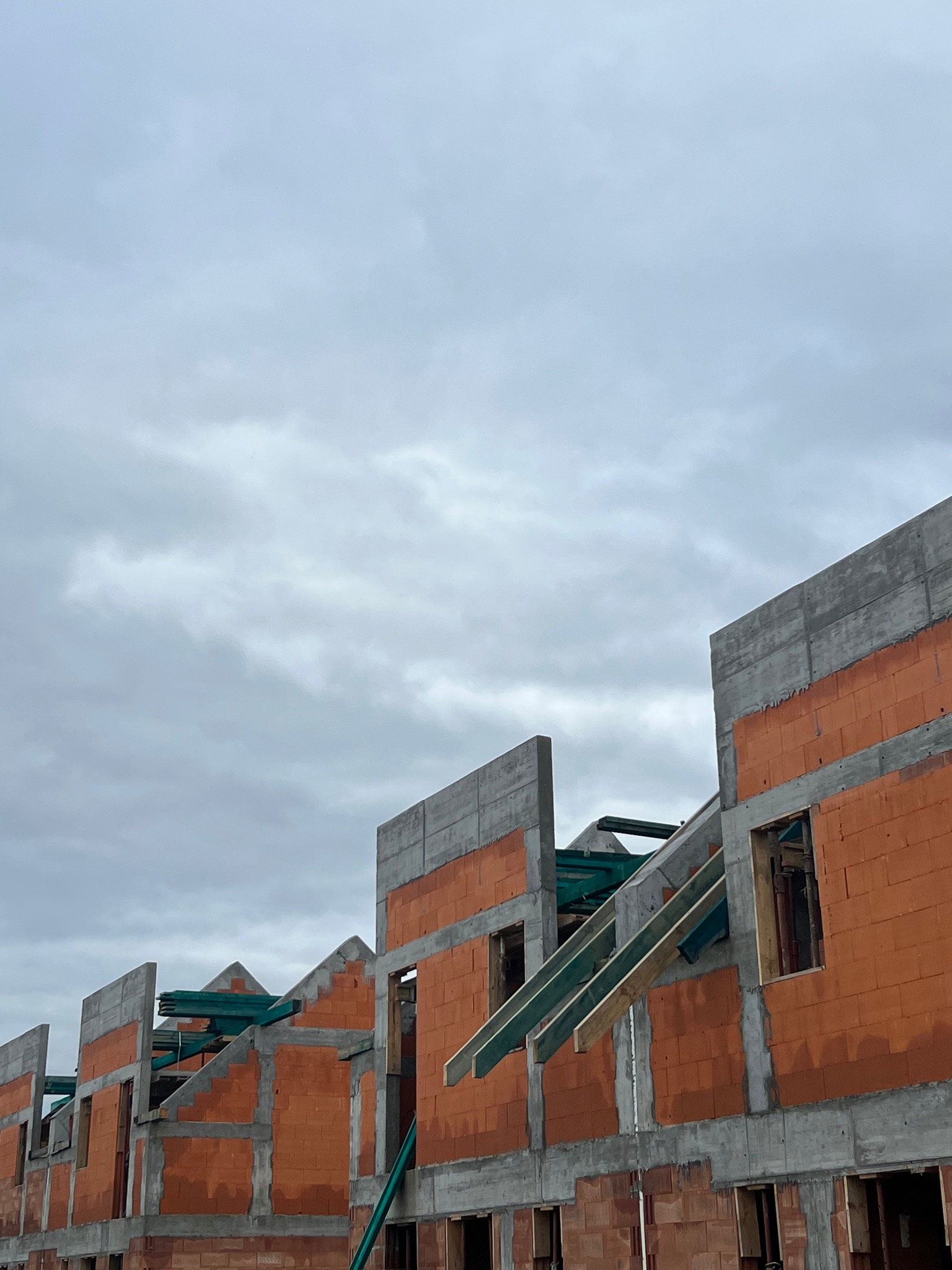 Surowy stan budowy domów z czerwonej cegły i betonu pod pochmurnym niebem. Widoczne elementy konstrukcyjne i otwory okienne.