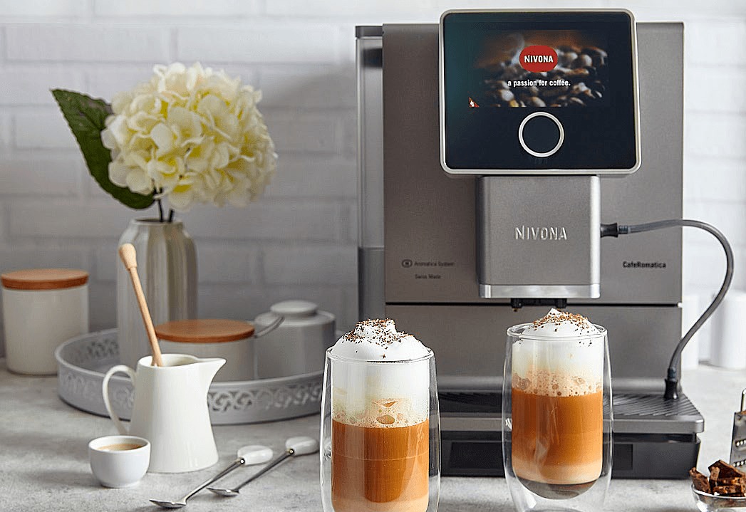Szary ekspres automatyczny Nivona CafeRomatica z dwoma szklankami latte macchiato z bitą śmietaną i posypką czekoladową na blacie, w tle wazon z białymi kwiatami hortensji i akcesoria do kawy.