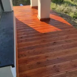 Wykończony taras z desek kompozytowych w ciepłym odcieniu brązu, z widocznymi słupami podtrzymującymi zadaszenie i fragmentem okna po lewej stronie.