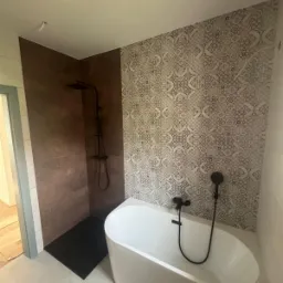 Nowoczesna łazienka po remoncie: biała wanna wolnostojąca, czarna armatura, płytki dekoracyjne na ścianie i brązowe płytki w strefie prysznicowej z odpływem liniowym.