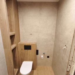 WykończeniaPro WOJCIECH JURASZEK - Nowoczesna łazienka z podwieszaną toaletą, drewnopodobne płytki na podłodze i ścianach, zabudowa z półkami. Minimalistyczny design, ciepłe odcienie.