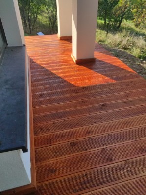 Wykończony taras z desek kompozytowych w ciepłym odcieniu brązu, z widocznymi słupami podtrzymującymi zadaszenie i fragmentem okna po lewej stronie.